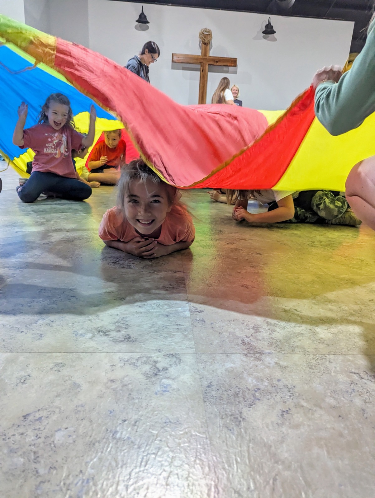 Children playing with a colorful rainbow parachute in the fellowship hall, one smiling child lying on the floor beneath it