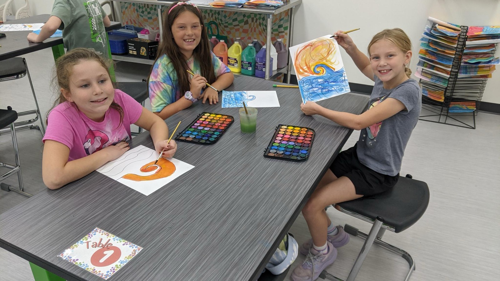 Three students painting with watercolors in art class, one holding up her finished sunrise painting