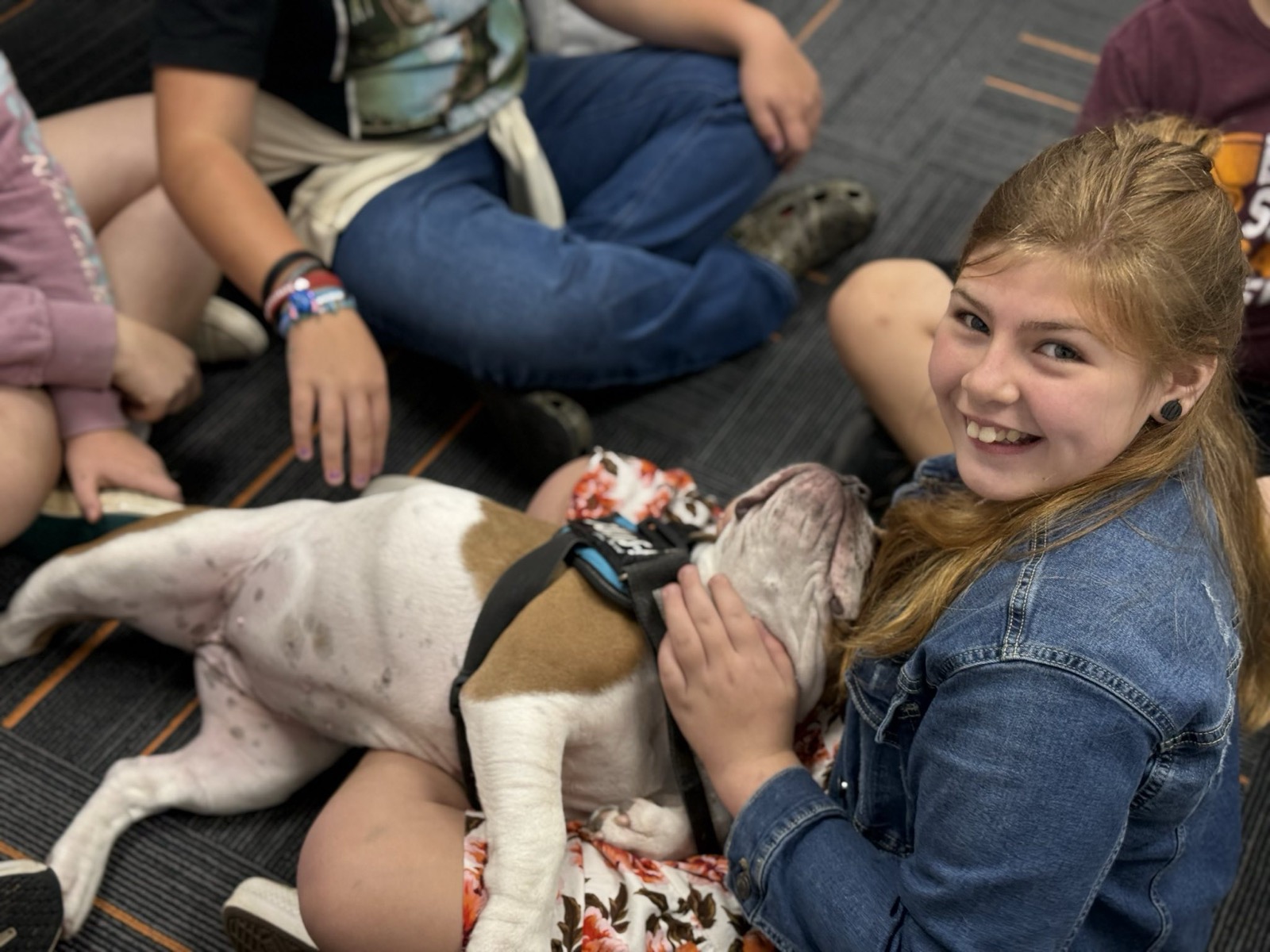 A smiling student in a denim jacket pets a bulldog wearing a service harness while classmates sit together on the floor