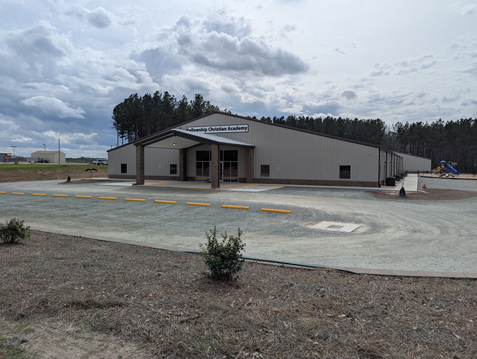 The Fellowship Christian Academy campus: a single-story building with a covered brick entrance, a playground to one side, and pine trees under a cloudy sky