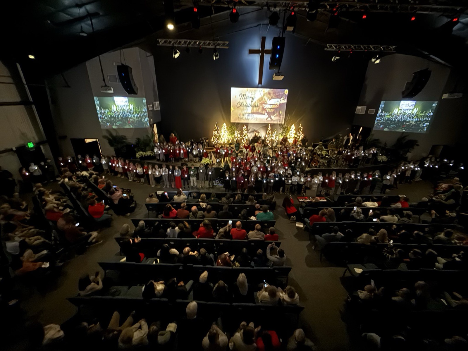 Students on stage holding lights during the FCA Christmas program, with a lit cross and families watching from the auditorium