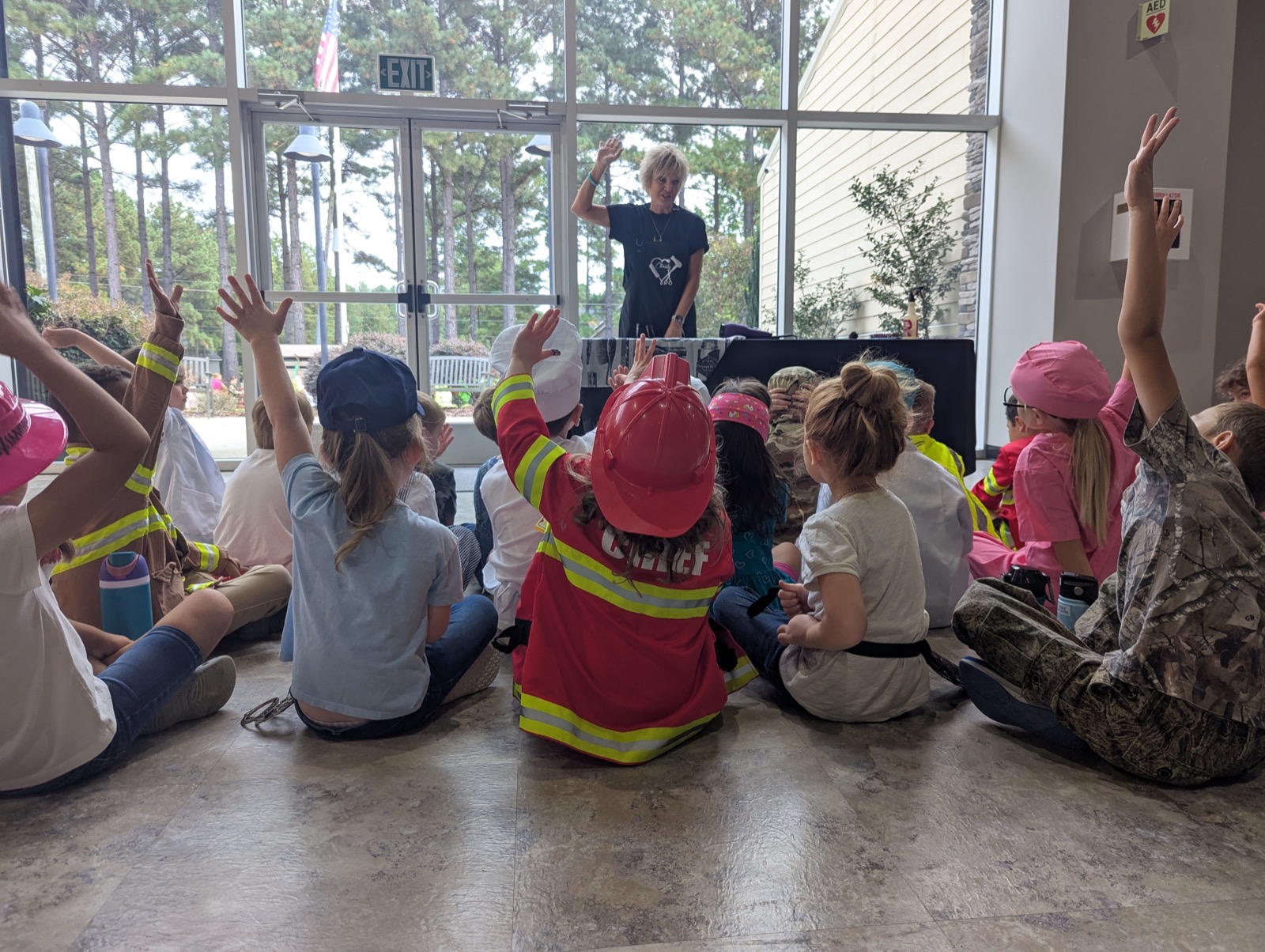 Students seated on the floor with hands raised during a Community Helpers lesson, some in firefighter dress-up, as a teacher leads at the front